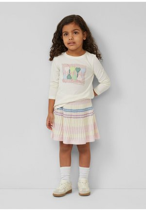 Junges Mädchen mit lockigem Haar, das ein langärmliges Hemd mit Herzen und einen gestreiften, plissierten Rock, weiße Socken und cremefarbene Turnschuhe trägt.