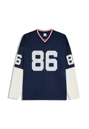 Tmavomodrý športový dres s bielym sieťkovým číslom 86 na prednej strane a rukávoch, vrstvený cez dlhé biele rukávy, s červeným lemovaním na výstrihu do V.