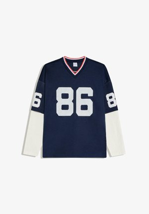 Marineblå sportstrøje med hvidt meshnummer 86 på front og ærmer, lagdelt over lange hvide ærmer, med rødkanter på V-halsen.