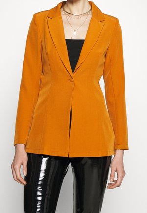 Kvinde iført en tætsiddende orange blazer over en sort top, kombineret med skinnende sorte bukser og lagdelte gyldne halskæder, stående mod en hvid baggrund.