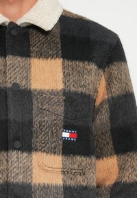 Black and tan plaid shirt jacket made of fluffy fabric, featuring a cream collar and a small "TOMMY JEANS" logo on the left pocket.