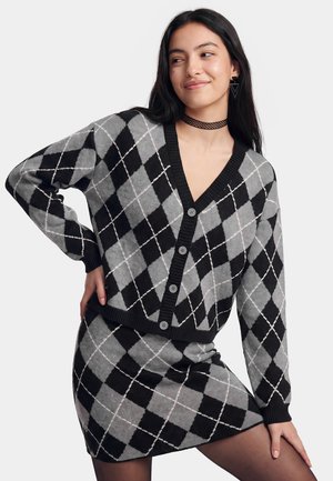 Vrouw met een grijs-zwarte argyle gebreide cardigan en bijpassende rok, poseert met één hand op de heup en glimlacht licht naar de zijkant.