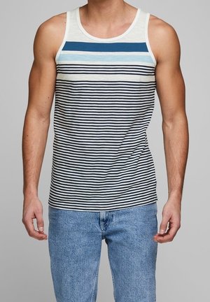 Mand iført ærmeløs tanktop med vandrette blå, lyseblå og sorte striber, kombineret med lyseblå denimjeans, stående mod en grå baggrund.