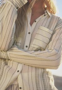 Chemise boutonnée avec une base crème à rayures noires, une poche avant et un tissu texturé. Coupe douce et décontractée avec des accents de boutons.