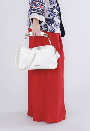 Donna che tiene una borsa a mano in pelle bianca con dettagli dorati, indossa una gonna rossa e una maglia con motivo blu e bianco.