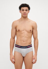 Pilkos medvilnės apatiniai su tamsiai mėlynu juosmeniu, puoštas raudonomis ir baltomis juostelėmis bei Lacoste logotipu priekyje. Glotni tekstūra.