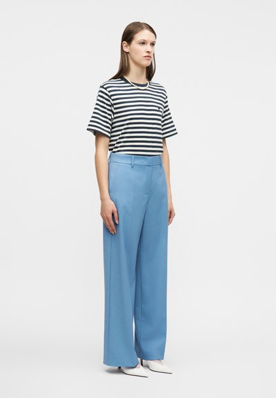 Γυναίκα που στέκεται, φοράει ένα κοντομάνικο πουκάμισο με ασπρόμαυρες ρίγες, ανοιχτό μπλε παντελόνι με φαρδύ πόδι, λευκές μυτερές γόβες και ένα κολιέ με μαργαριτάρια.