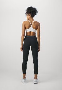 White sports bra and black leggings with a high waistband, featuring Nike branding. Model is standing with back to camera, wearing white sneakers.