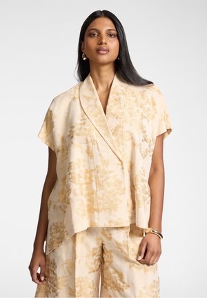 Donna che indossa una giacca a maniche corte color beige con motivo floreale e pantaloni larghi abbinati, accessoriata con orecchini a cerchio dorati e braccialetto.