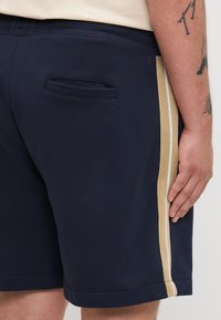 Marinblå shorts med en slät yta, med en sidostripa i guld och vitt. Inkluderar en bakficka.
