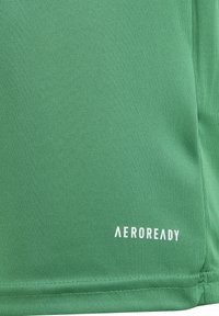 Primer plano de tela verde con la marca "AEROREADY" en blanco cerca del dobladillo inferior, mostrando una fina textura de punto y detalles de costura.