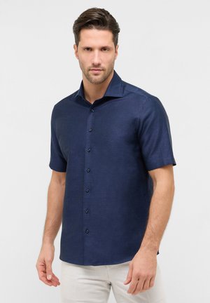 Mand iført en mørkeblå, kortærmet skjorte med knapper og lyse beige bukser, stående mod en ensfarvet lys baggrund.