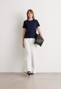 Femme marchant à l'intérieur, portant un t-shirt bleu marine, un pantalon blanc à jambes larges, des chaussures noires et tenant un sac à main en cuir noir.