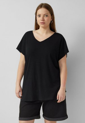 Jeune femme aux cheveux châtain clair portant un haut noir ample à col en V et un short noir retroussé, debout devant un fond clair uni.