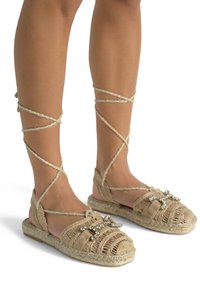 Płaskie sandały z plecionym wzorem z juty, w kolorze brązowym, z krzyżującymi się wiązaniami wokół kostki oraz dekoracyjnymi koralikami na palcach.
