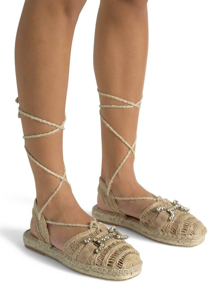 Płaskie sandały z plecionym wzorem z juty, w kolorze brązowym, z krzyżującymi się wiązaniami wokół kostki oraz dekoracyjnymi koralikami na palcach.
