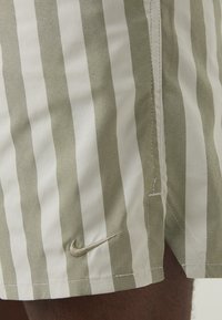 Nike swim shorts with vertical olive green and white stripes, featuring a small gold logo and lightweight fabric. Stitched seams are visible.