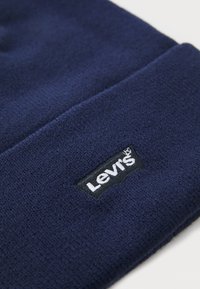 Tamsiai mėlynas megztas šalmukas su juodu Levi's logotipu, kuriame balta tekstas ant apvado.