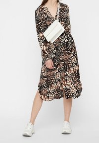 Abito lungo nero e marrone a fantasia con scollatura a V, maniche lunghe e spacco davanti. Abbinato a una borsa a tracolla bianca e sneakers bianche.