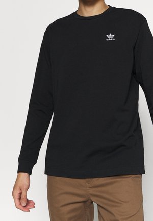 Person wearing a black long-sleeve Adidas shirt with white logo on chest and brown pants, standing against a plain light background.