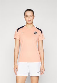 Maillot de sport pêche avec des accents bleu marine, col rond et manches courtes. Présente le logo de Chelsea FC et le swoosh de Nike en or sur la poitrine.