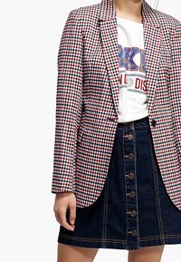 Czerwona i biała marynarka w kratę z wycięciem na klapach, noszona na białym t-shircie z nadrukiem, zestawiona z niebieską jeansową spódnicą z guzikami z przodu.