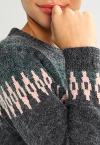 Suéter de punto en gris oscuro con un patrón geométrico en teal y rosa. Presenta un tejido texturizado y puños acanalados, resaltando diseños en capas.