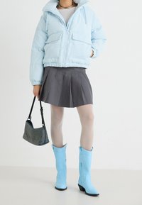 Hellblaue Cord-Pufferjacke mit Reißverschluss, grauer plissierter Rock, durchsichtige Strumpfhosen und blaue Cowboy-Stiefel. Schwarze und silberne Handtasche inklusive.