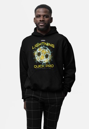 Homme portant un sweat à capuche noir avec un ballon de football lumineux et le texte jaune « Official Lightning Quick Pro » ainsi qu’un pantalon noir avec un motif en grille blanche.