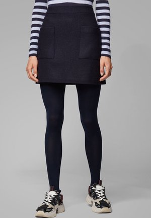 Laivastonsininen villasekoitehame, jossa on kaksi etutaskua, puettuna laivastonsinisten läpinäkymättömien sukkahousujen ja mustien valkoisin yksityiskohdin varustettujen tennarien päälle. Raidallinen neule näkyy yläpuolella.