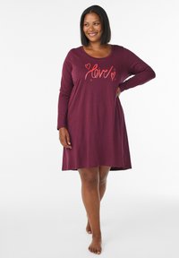 Donna sorridente in piedi a piedi nudi, che indossa un abito a maniche lunghe color bordeaux con la parola "love" e disegni di cuori rossi sul petto.