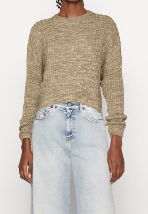 Personne portant un pull à manches longues beige texturé et un jean large taille haute bleu clair sur un fond uni.