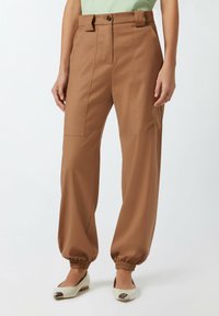 Smeđe hlače širokih nogavica od glatkog materijala, s kopčanjem na dugme, petljama za remen i elastičnim manžetama. Uparene s bijelim balerinkama.