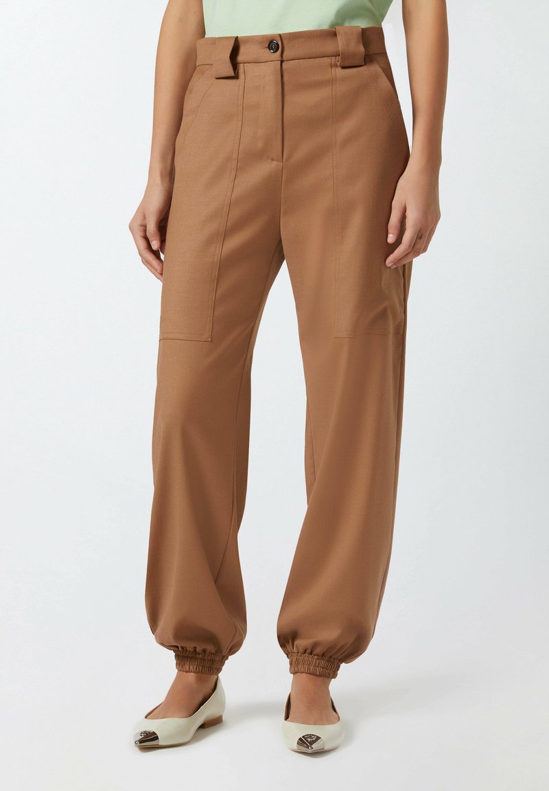 Smeđe hlače širokih nogavica od glatkog materijala, s kopčanjem na dugme, petljama za remen i elastičnim manžetama. Uparene s bijelim balerinkama.