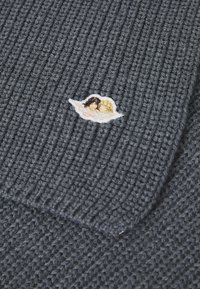 Strikket mørkegrå sweaterstof med en lille broderet patch af to figurer med sort og blond hår, sat mod en hvid sky-kontur.