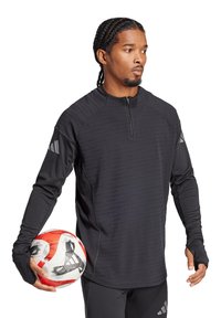 Maglietta sportiva nera a maniche lunghe con colletto a mezza zip, tessuto testurizzato e aperture per il pollice, abbinata a un pallone da calcio rosso e bianco.