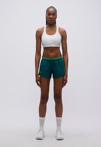 Αθλητική γυναίκα στέκεται ακίνητη φορώντας λευκό αθλητικό σουτιέν, πράσινα σορτς Under Armour, λευκές κάλτσες και λευκά αθλητικά παπούτσια σε απλό φόντο.