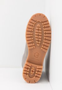 Semelle d'une botte beige avec des crampons profonds et texturés, positionnée verticalement contre des surfaces blanches, affichant le nom de la marque et les détails de la taille.