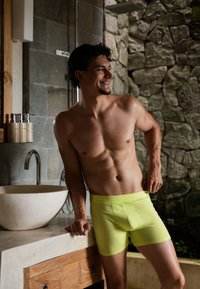 Neon gele boxershorts met een zachte textuur en een contrasterende witte tailleband met het logo "HAPPY SHORTS", gepresenteerd tegen een achtergrond van natuurlijke stenen.