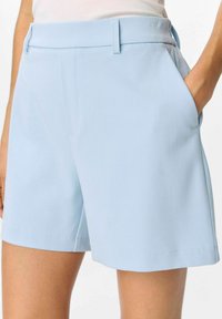 Pantaloni corti azzurri su misura realizzati in tessuto morbido con tasche laterali, passanti per la cintura e un orlo pulito. Presentano una vestibilità classica e dettagli minimali.