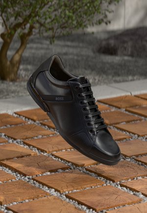Baskets en cuir noir avec des accents texturés, surface lisse et design à lacets. Présente un logo sur le côté. Élevées au-dessus d'un sol à motifs.