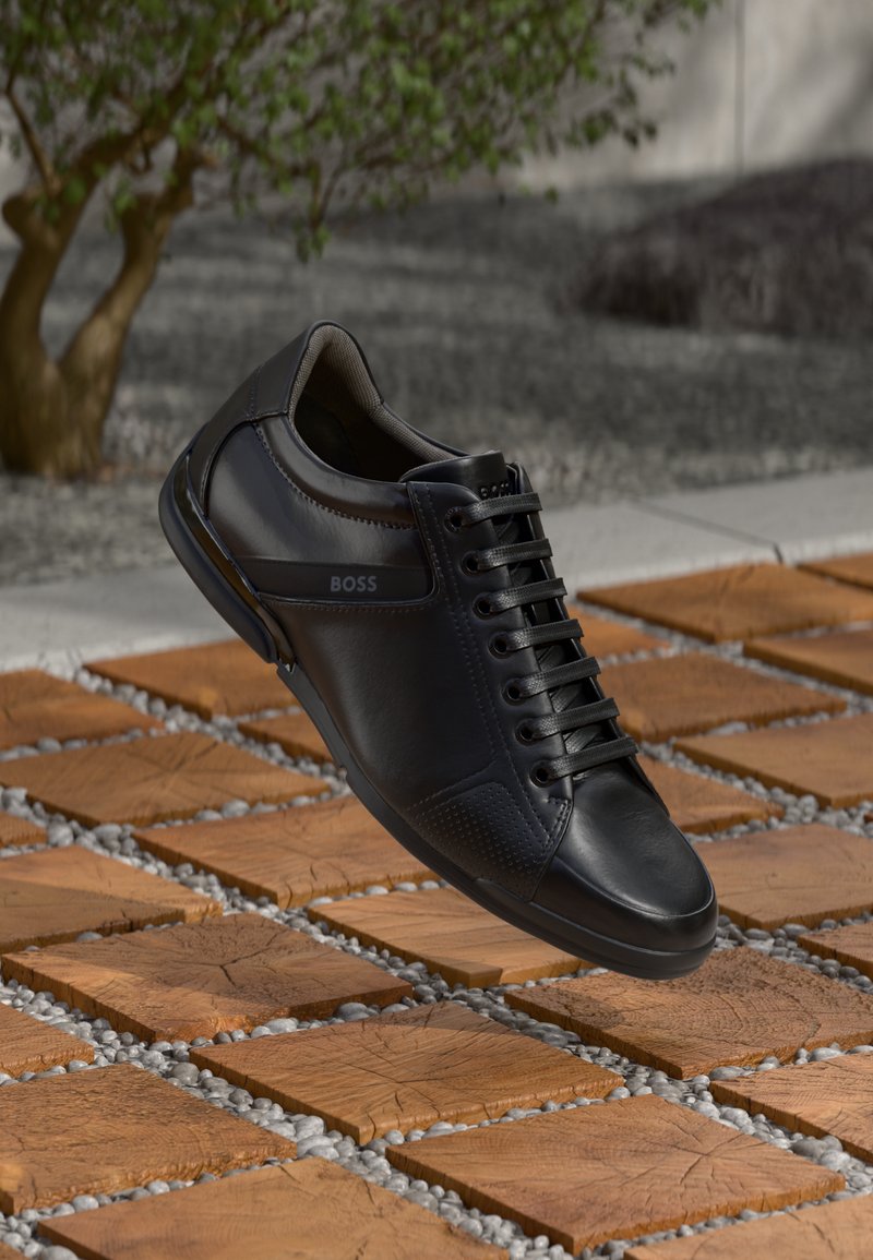 Zwarte leren sneaker met getextureerde accenten, een gladde oppervlakte en een vetersluiting. Voorzien van een logo aan de zijkant. Geplaatst boven een gepatternde ondergrond.