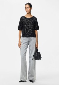 Mulher vestindo uma blusa preta de lantejoulas de manga curta, jeans claros de pernas largas, sapatos pretos de bico fino e segurando uma pequena bolsa preta.