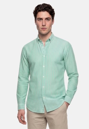 Hombre vestido con una camisa de botones de rayas verticales verdes y blancas y pantalones beige, de pie con una mano en el bolsillo.