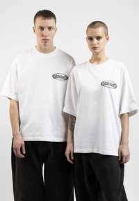 White oversized t-shirts with a black logo on the chest, paired with loose black denim pants. Smooth cotton texture, relaxed fit.