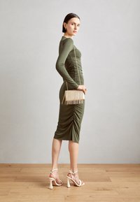 Sukienka w kolorze oliwkowej zieleni, marszczona, z długimi rękawami, do kolan. Uzupełniona torebką z złotym łańcuszkiem i beżowymi sandałami na wysokim obcasie.