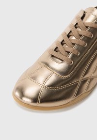 Metallic gold sneaker featuring a smooth leather upper, lace closure, and textured stitching details. Round toe and flexible rubber sole.