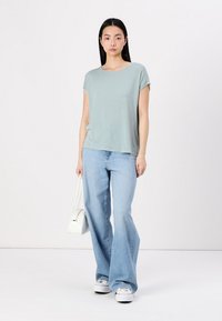 Lyseblå T-shirt med rund hals og korte ærmer, parret med lysvaskede flared jeans og hvide sneakers. Model, der holder en lille hvid taske.