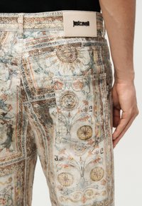Ανδρικά τζιν με περίπλοκα μοτίβα ήλιου, λουλουδιών και προσώπου λιονταριού σε απαλούς παστέλ τόνους, με μπεζ ετικέτα επωνυμίας Just Cavalli και πίσω τσέπη.
