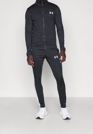 Mężczyzna ubrany w czarny dres Under Armour z białymi paskami po bokach i białe sportowe buty, stojący na jasnym, jednolitym tle.